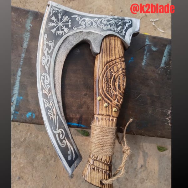 Bearded Viking axe: A deeply curved bearded head with contrasting scrollwork and a compass motif. The carved wooden handle has a sculpted grip, cord wrapping and a hanging loop.