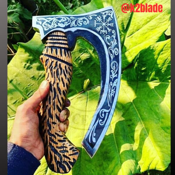 Norse hatchet: An elongated bearded head with bright scrollwork and a compass motif against a dark background. The short wooden handle features bold carving and a rounded grip.